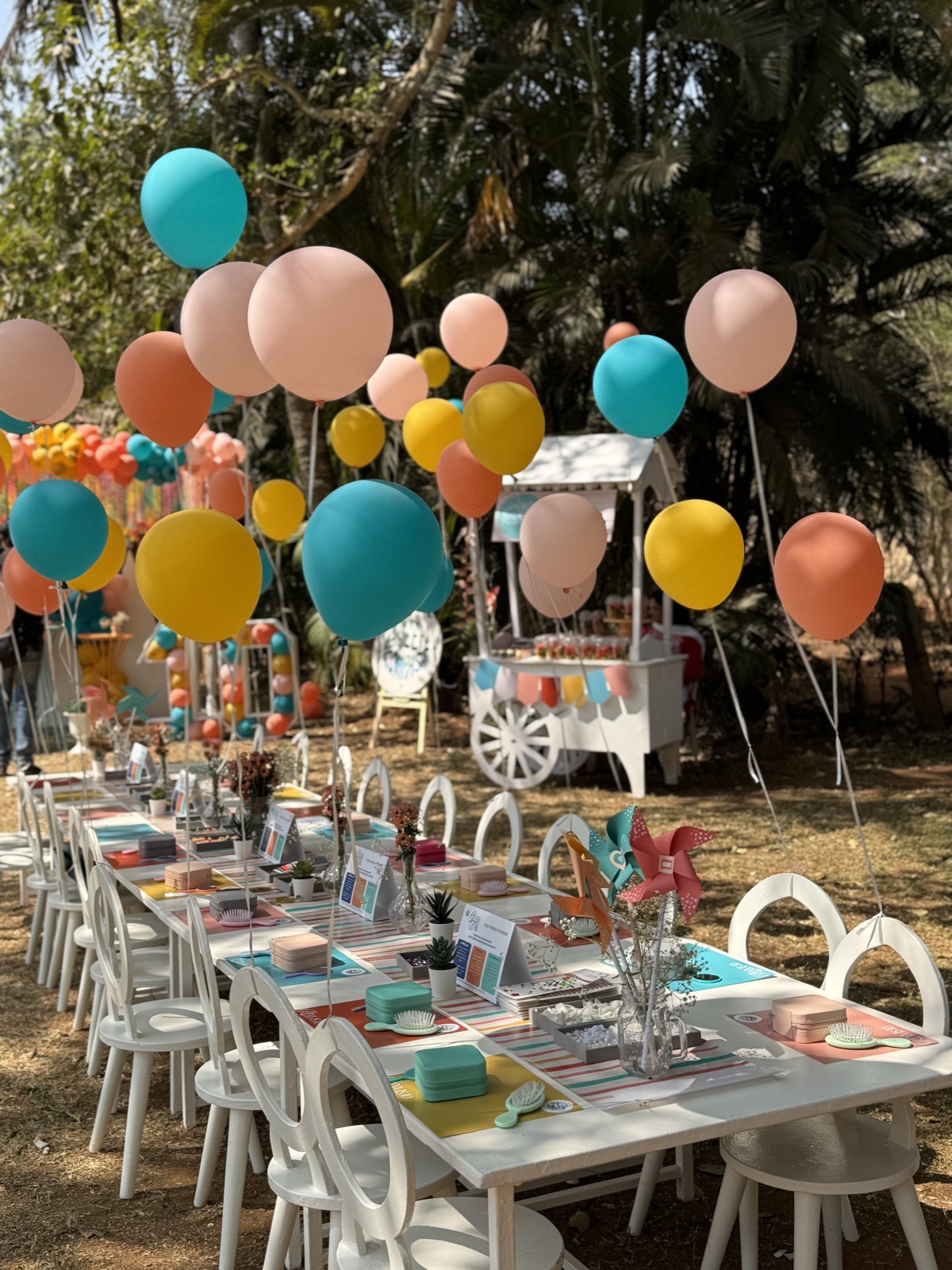 A long white table set under trees for a decorating activity, with balloons on strings above every place and a sweet cart at the far end